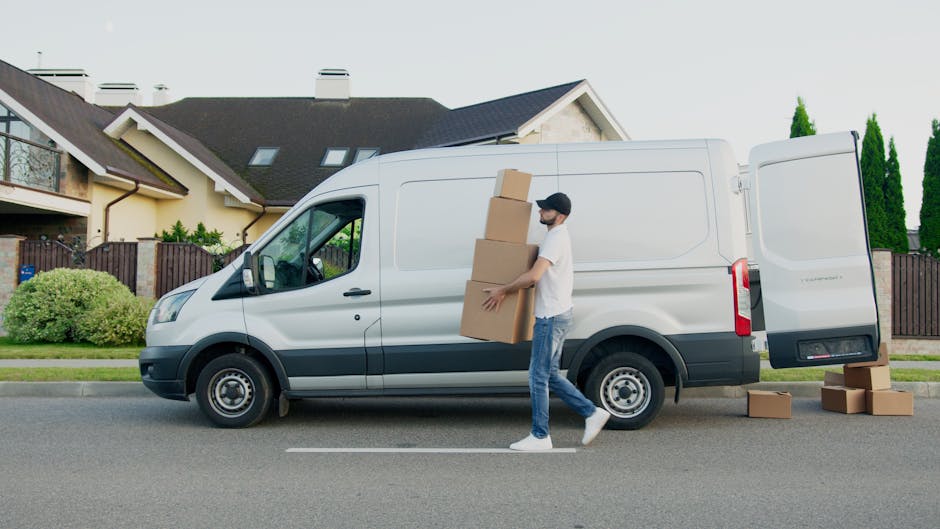 A man dressed in a blue jacket and blue trousers is actively loading a cardboard box into the rear cargo area of a white moving van parked on a cobbled street. The van's rear doors are open, revealing