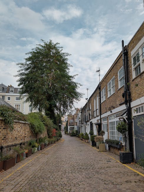 A narrow cobblestone alleyway in Bayswater, lined with residential buildings on the right side featuring white-framed windows and light brown brick facades, with a row of potted plants at the base of the buildings. On the left side, a brick wall and greenery are visible, including a tall tree with dense foliage extending above the wall. The sky is partly cloudy with patches of blue. This setting depicts an outdoor area where home relocation or furniture transport activities might occur, aligning with the services offered by Man and Van Bayswater, such as house removals and moving logistics.