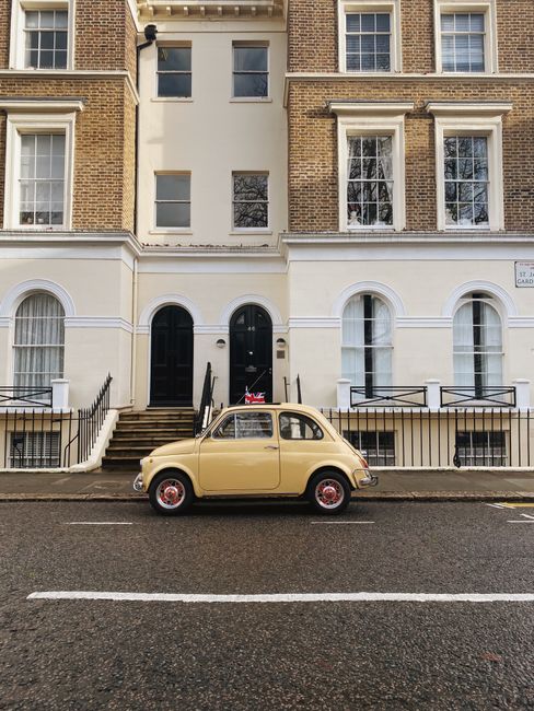 A beige vintage-style car parked on the street directly in front of a multi-storey residential building with a brick façade and white decorative elements around large windows. The building has steps