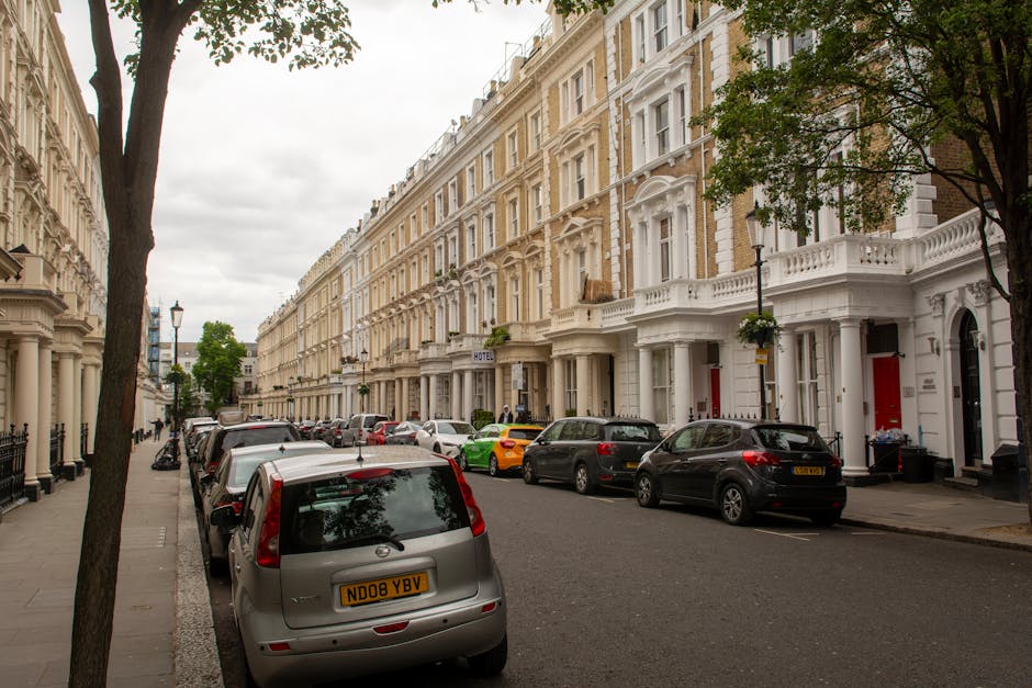 Image showing a residential street with a row of white Victorian-style terraced houses, each featuring ornate architectural details and bay windows. Several parked cars, including a silver Mini with a