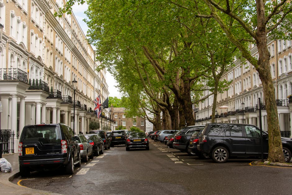 Image showing a residential street with a row of white Victorian-style terraced houses, each featuring ornate architectural details and bay windows. Several parked cars, including a silver Mini with a